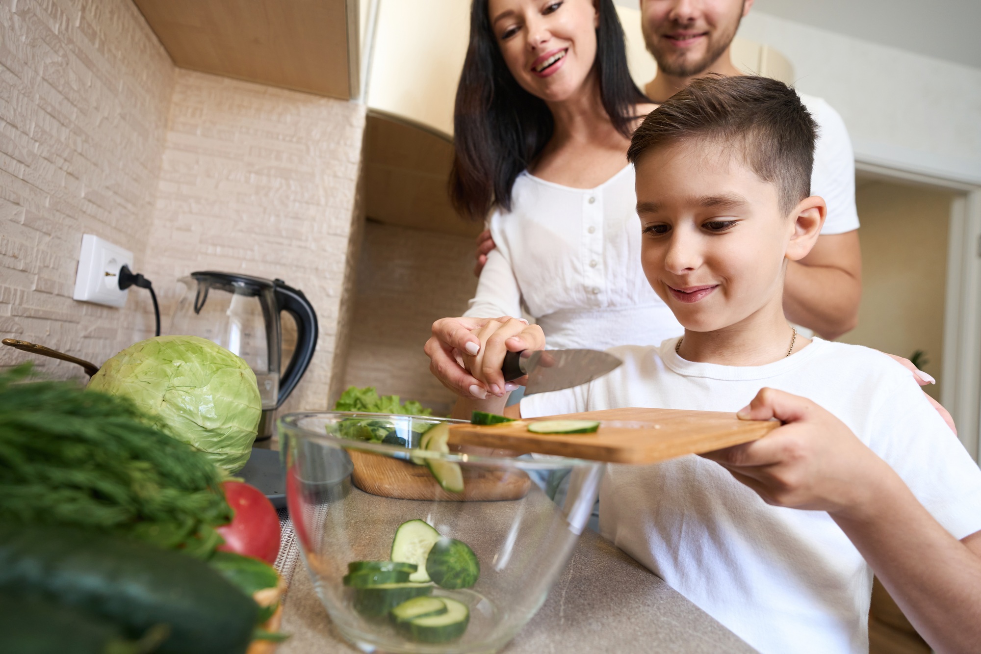 Mère et père debout derrière le garçon et en regardant comment il pousse des morceaux de concombre dans le bol de la planche à découper
